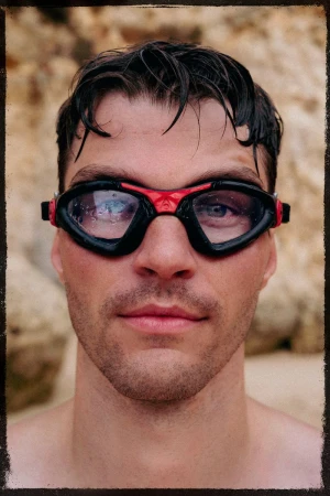 Close-up portrait of a man wearing black and red swimming goggles with water droplets, against a blurred sandy background.