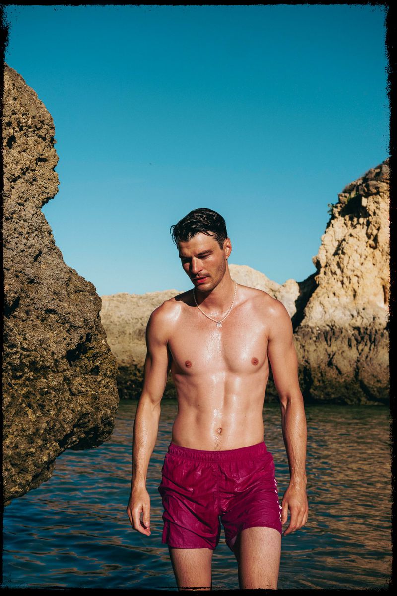 A muscular man in magenta swim trunks stands in shallow water, flanked by rocks under a clear blue sky in a sunlit outdoor setting.