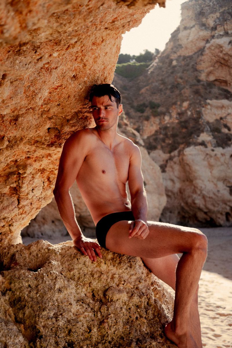 Man in swimsuit posing by beach rocks during daytime. Tanned skin and dark hair are visible. Rocky beach and cliffside setting.