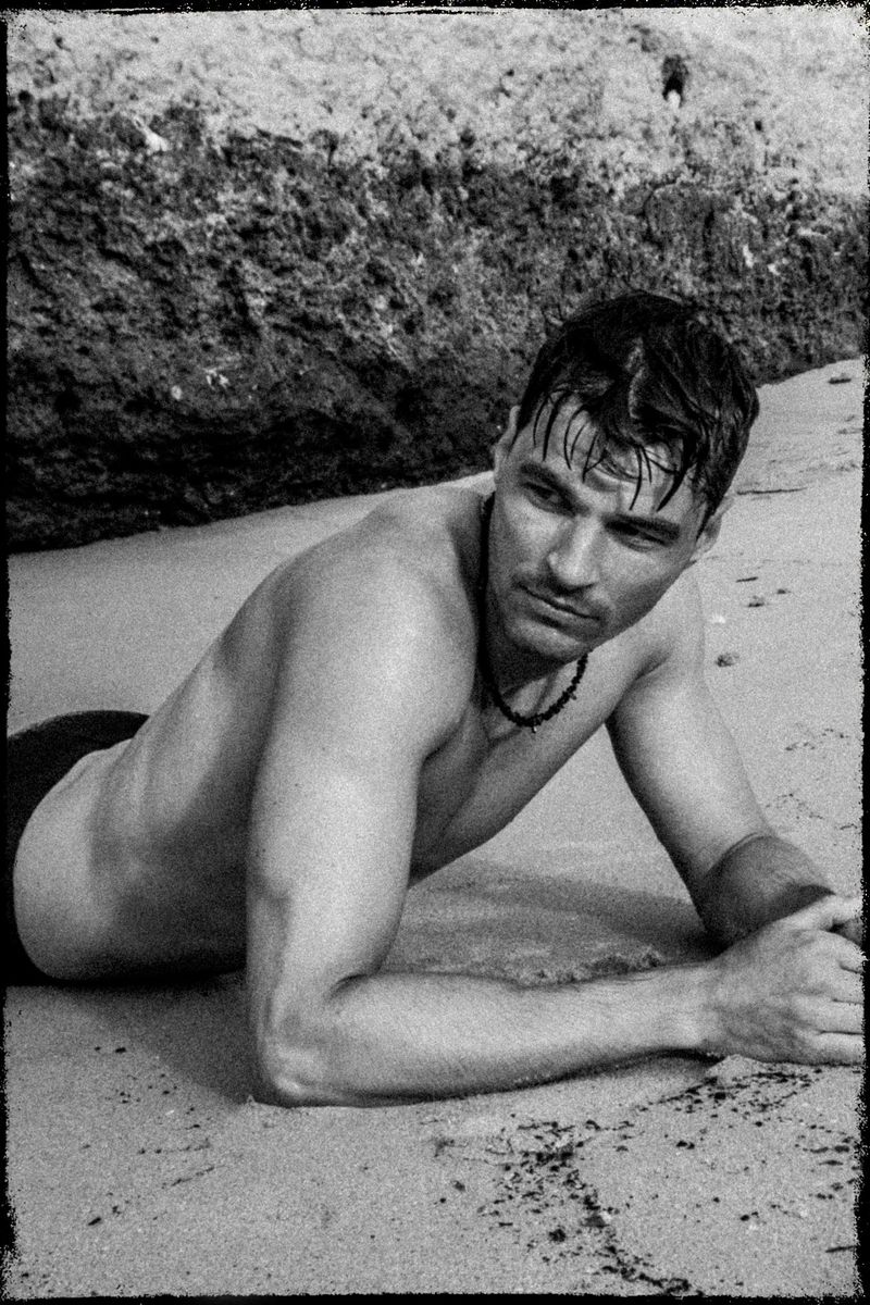 Black-and-white portrait of an adult male lying on a beach with a rocky background, photographed in an editorial style.