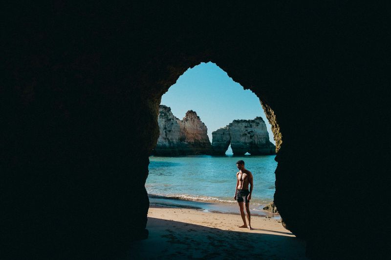 Model Arran Sly standing on a sandy beach in dark swim shorts, framed by the silhouette of a natural sea cave archway.