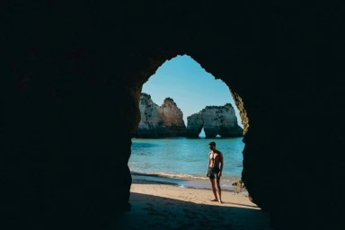 Model Arran Sly standing on a sandy beach in dark swim shorts, framed by the silhouette of a natural sea cave archway.