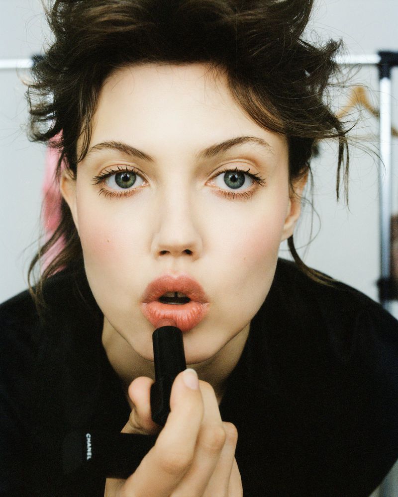 Close-up of a young woman applying dark lipstick in a photo studio, wearing a black top, and with hair styled in loose waves.
