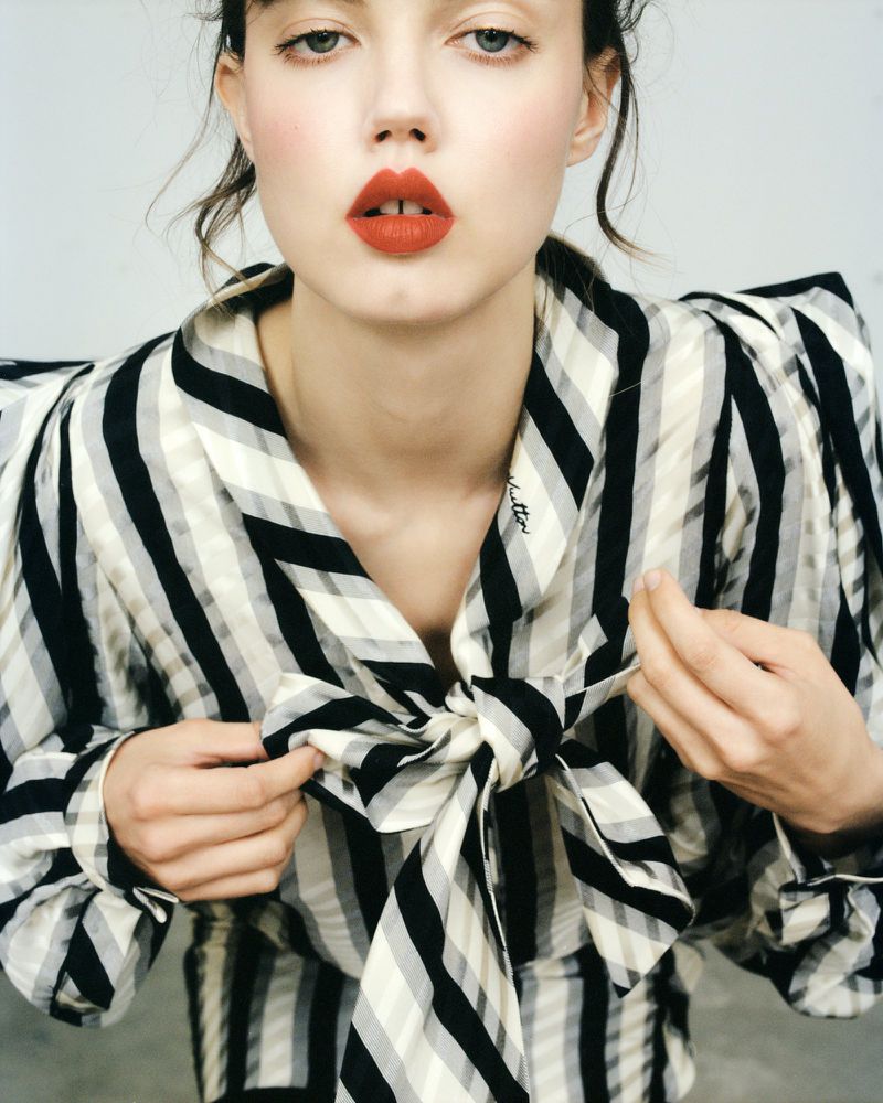 Portrait of a fair-skinned woman with red lipstick wearing a striped shirt and bow, looking directly at the camera in a studio setting.