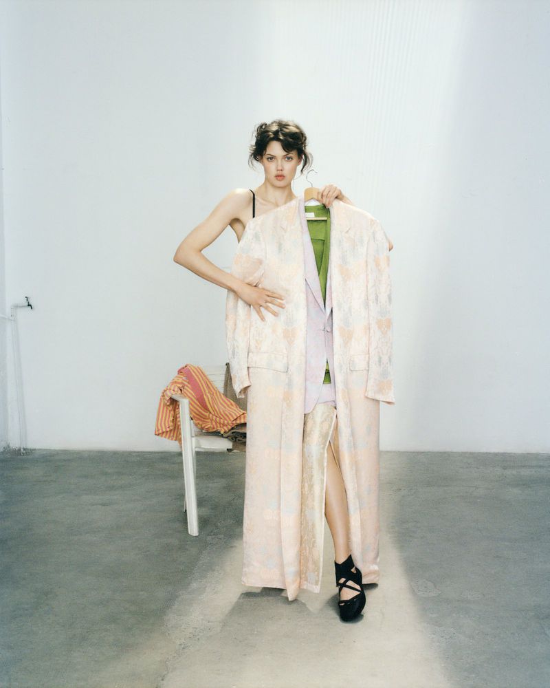 Brunette woman in platform heels poses in a photo studio while holding a jacket and robe with a patterned chair in the background.