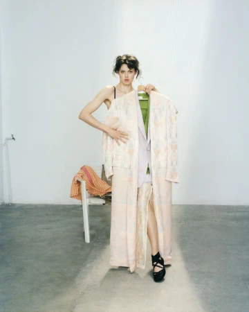Brunette woman in platform heels poses in a photo studio while holding a jacket and robe with a patterned chair in the background.