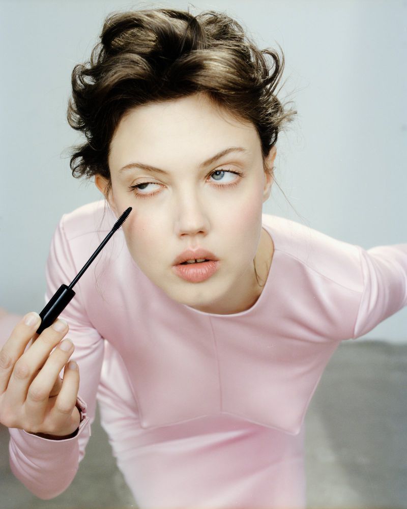 Young woman with dark hair applying mascara in a close-up shot, wearing a pink long-sleeved dress against a muted background.