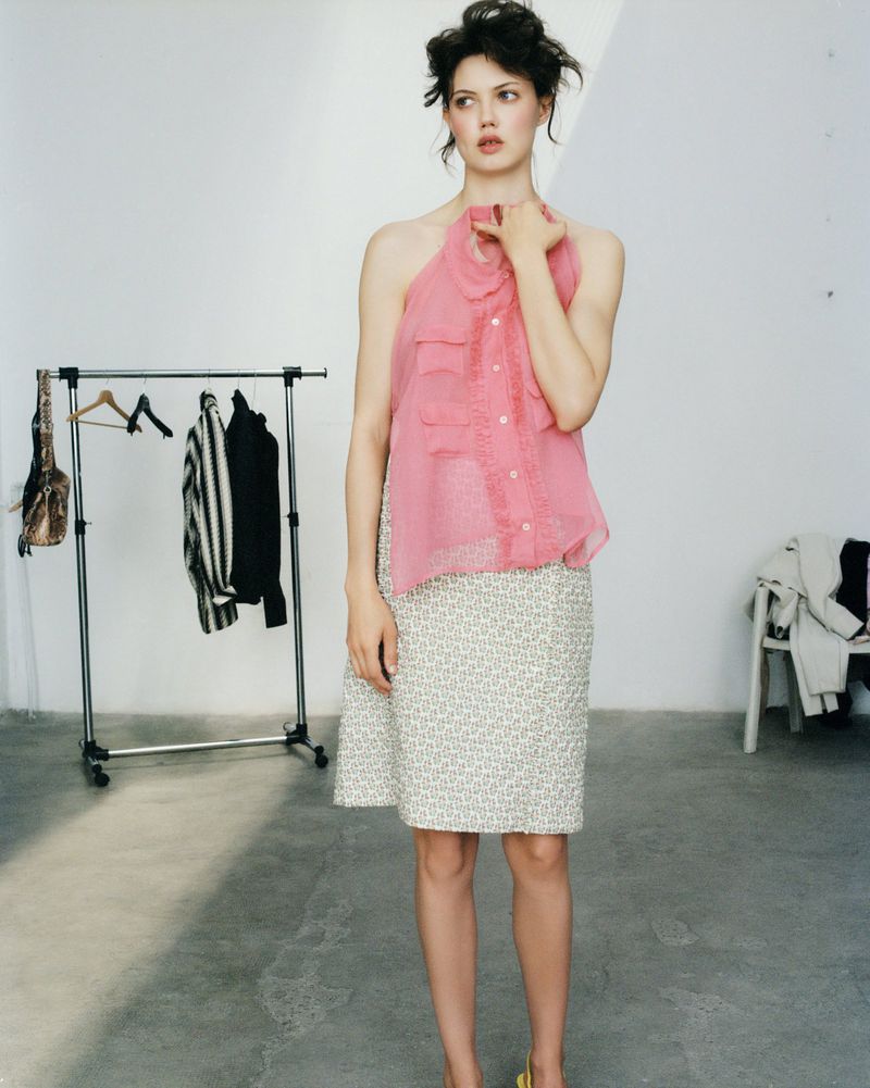 A woman in a pink halter top and patterned skirt stands in a photo studio, with a clothing rack and chair visible in the background.