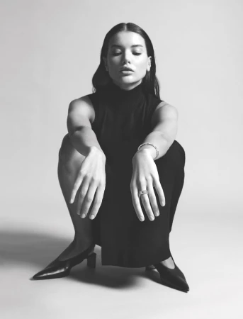Elegant woman in black dress poses seated with closed eyes. Black and white studio shot showcasing a contemporary fashion look.