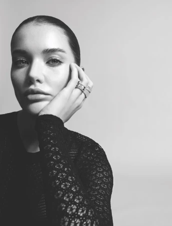 Monochrome portrait of a young adult female with her hand near her face, wearing several rings and a textured top in a studio setting.