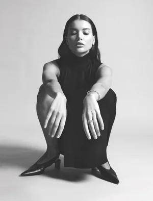Monochrome fashion editorial portrait of a woman crouching against a light grey background wearing a black sleeveless top.