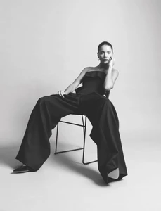 Monochrome fashion portrait of a woman seated on a minimalist chair wearing wide-leg black trousers and a strapless top.
