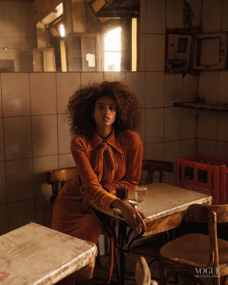 Woman with curly hair and an orange dress poses at a table with drinks in an indoor cafe setting, moody lighting, and Vogue magazine credit.