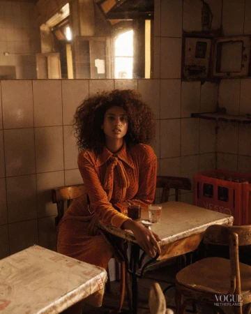 Woman with curly hair and an orange dress poses at a table with drinks in an indoor cafe setting, moody lighting, and Vogue magazine credit.