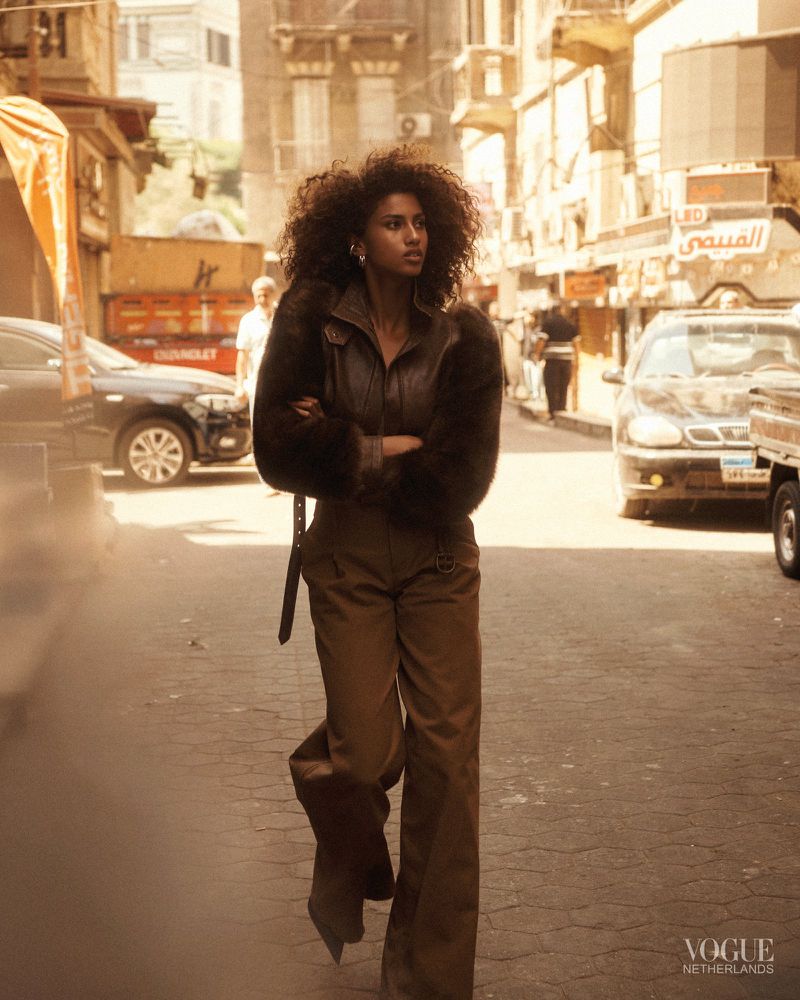 A woman with curly hair walks down a sunlit street in a brown fur-lined jacket and matching pants, exuding confidence in an urban setting.