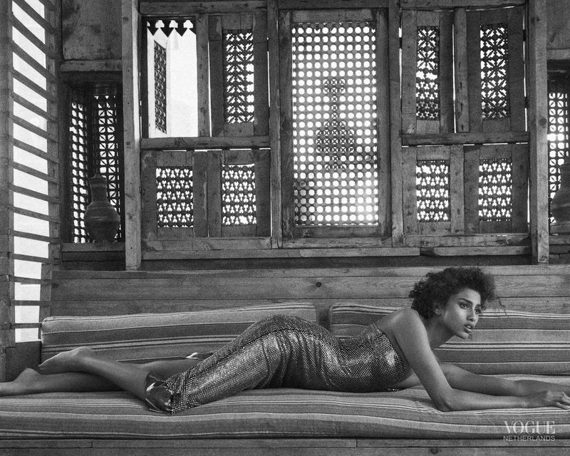 A black and white fashion shot of a curly-haired woman in a dress, reclining on a patterned surface in a room with ornate windows.
