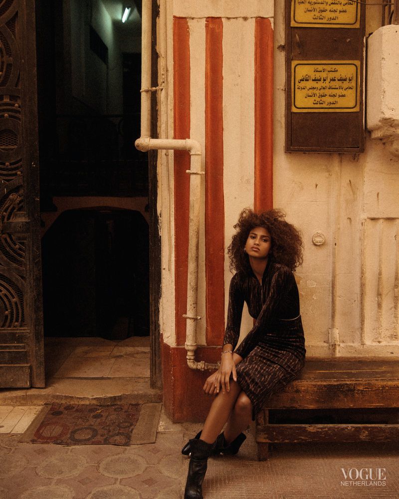 Woman with curly hair in vintage-style dress sits on bench near doorway with striped wall in urban setting, wearing boots.