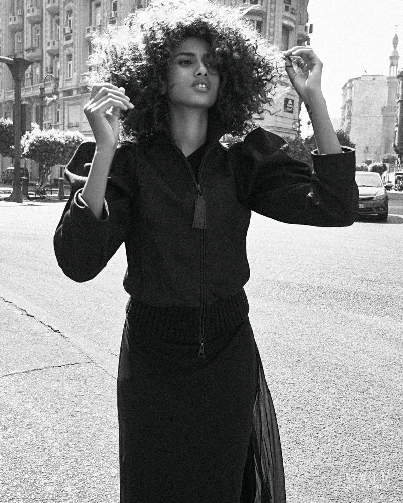 Black and white portrait of an adult female with afro hairstyle wearing a jacket and long skirt on a city street with buildings.