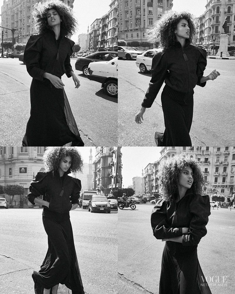 Black and white fashion shot of a woman with curly hair in a black outfit crossing a city street with buildings and cars in the background.