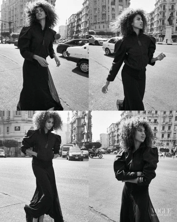 Black and white fashion shot of a woman with curly hair in a black outfit crossing a city street with buildings and cars in the background.