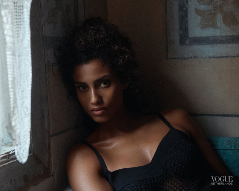 Close-up portrait of a woman with dark hair and tanned skin wearing a black mesh top, posing indoors near a window with white curtains.
