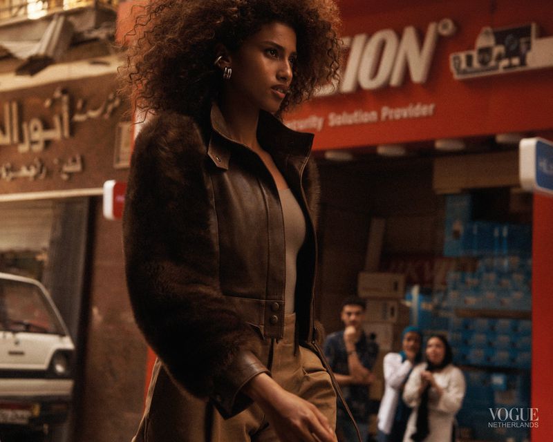 A woman with curly brown hair walks in an urban setting wearing a leather and fur jacket, with buildings and people in the background.