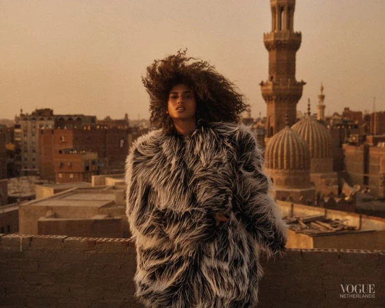 Woman with curly hair in fur coat on rooftop in Cairo, Egypt at sunset with minarets and buildings in background.