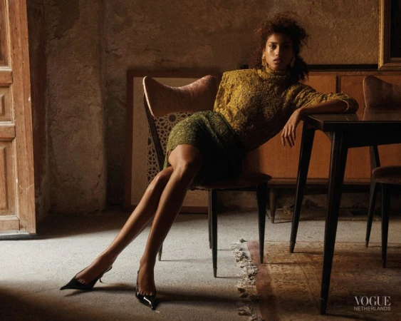 Model Imaan Hammam in an olive knit top and skirt seated on a chair beside a dark dining table in a warmly lit rustic room.