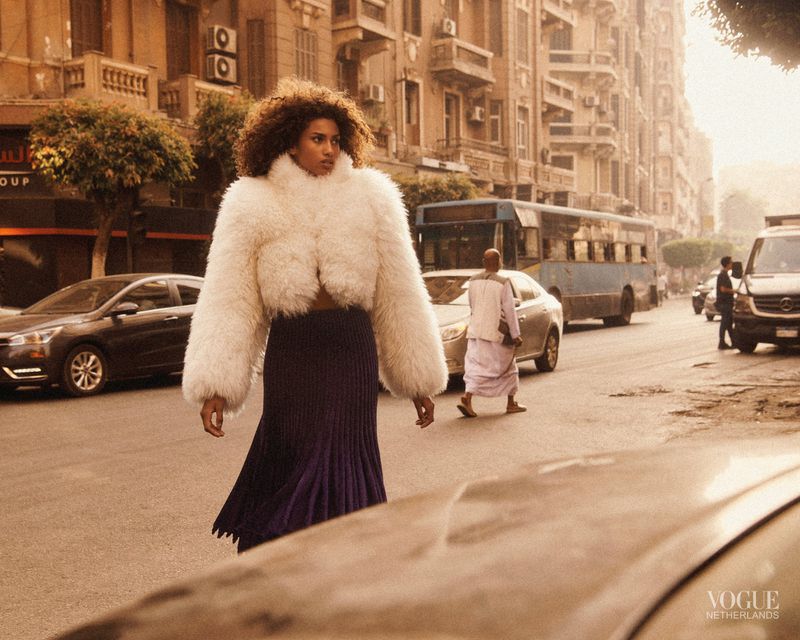 A model in a white jacket and purple skirt walks across a city street with cars and buildings in the background.