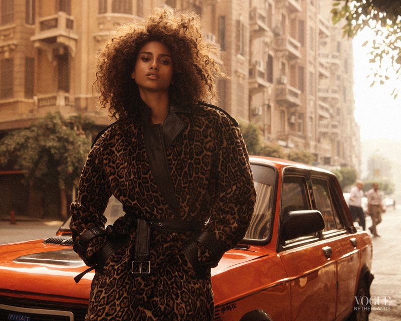 A stylish adult woman with curly hair poses in a leopard-print coat next to an orange Fiat in an urban scene, Vogue Netherlands logo in the corner.