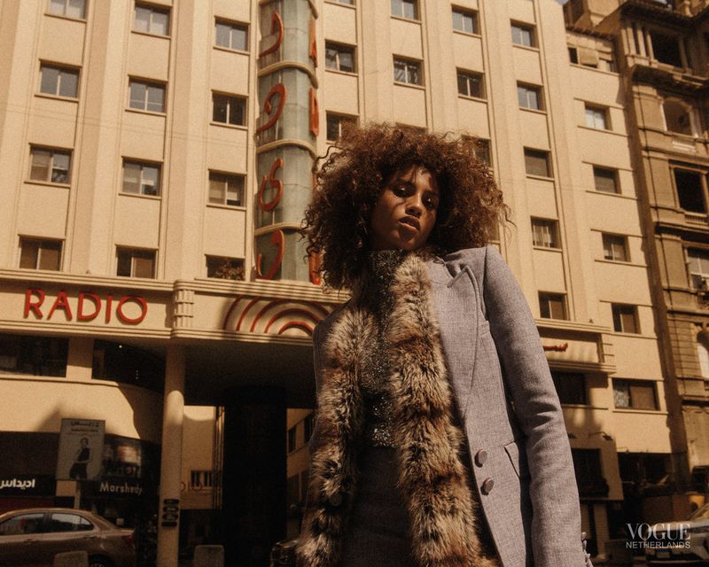Woman with curly hair wearing gray blazer with fur trim standing in front of building with 'RADIO' signage in a city setting.