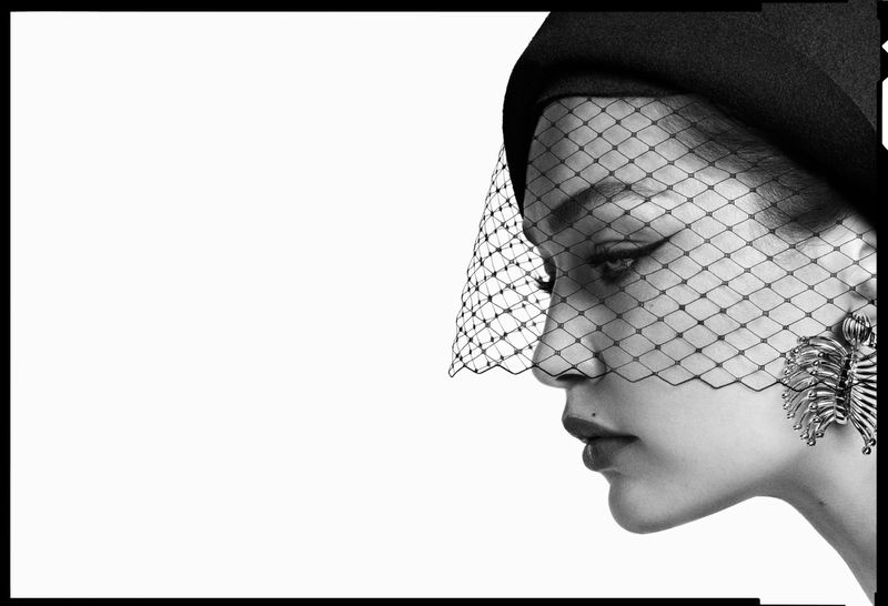 Monochrome profile of a woman wearing a black hat with veil and statement earrings against a bright white background in studio lighting.