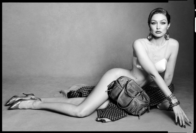 Black and white studio portrait of a reclining adult female fashion model wearing a bra and jewellery, with a handbag and high heels.