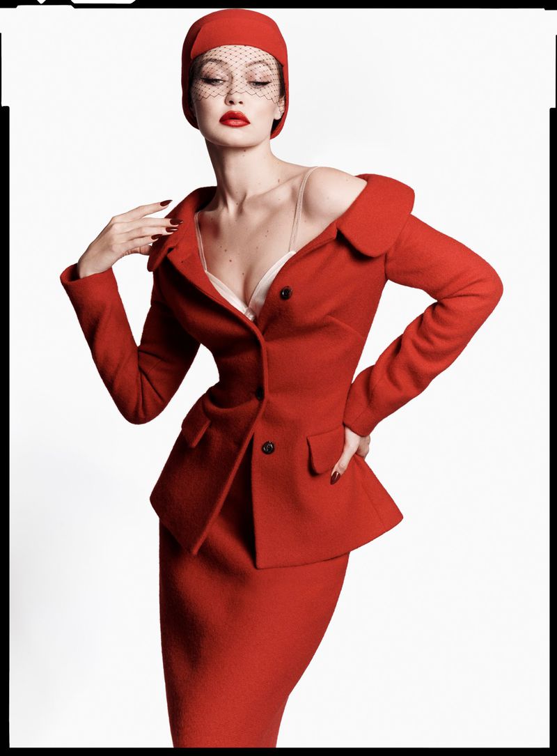 Woman in a red skirt suit and hat with net veil poses in a studio setting. Fashion editorial photography with clean backdrop.
