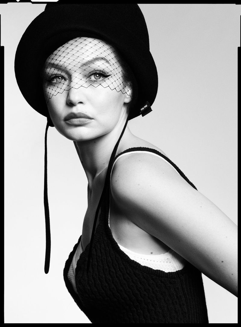 Portrait of a young woman with fair skin, wearing a hat with a net veil, and a dark tank top, set against a neutral background.