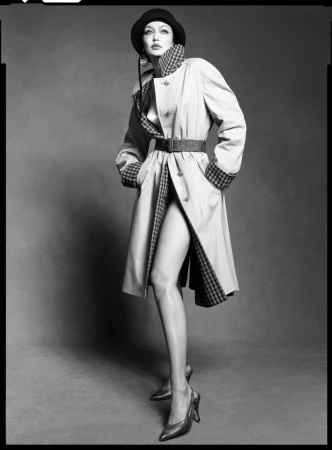 Gigi Hadid poses wearing a belted trench coat with checked lapels, a dark cloche hat, and heels against a grey studio backdrop.