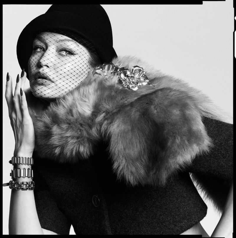 Black and white fashion portrait of a model wearing a hat with veil, fur collar, and jewelry, against a plain background.