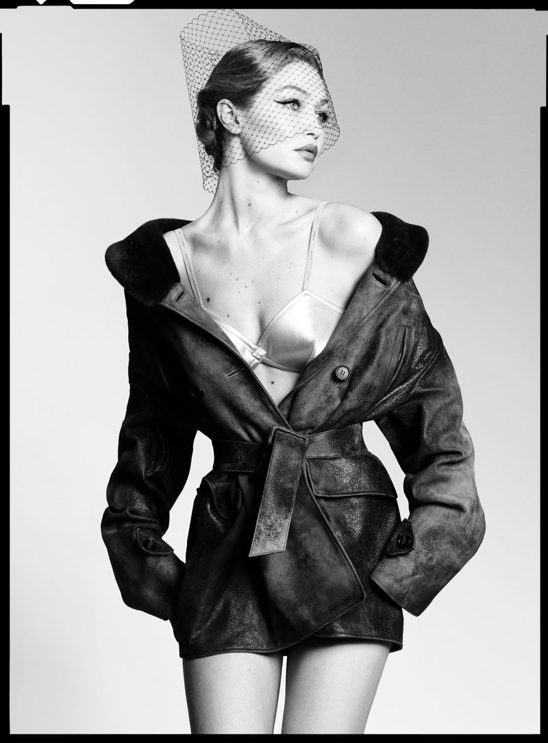 Black-and-white studio portrait of a woman wearing a net veil, unbuttoned leather jacket, and bra top, looking to the side.