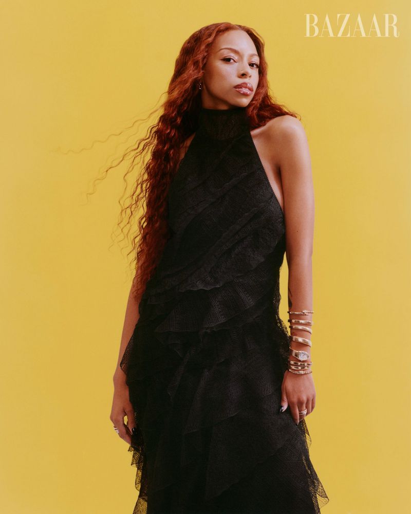 A woman with long red hair poses in a black halter dress against a yellow background, accessorized with multiple gold bracelets.