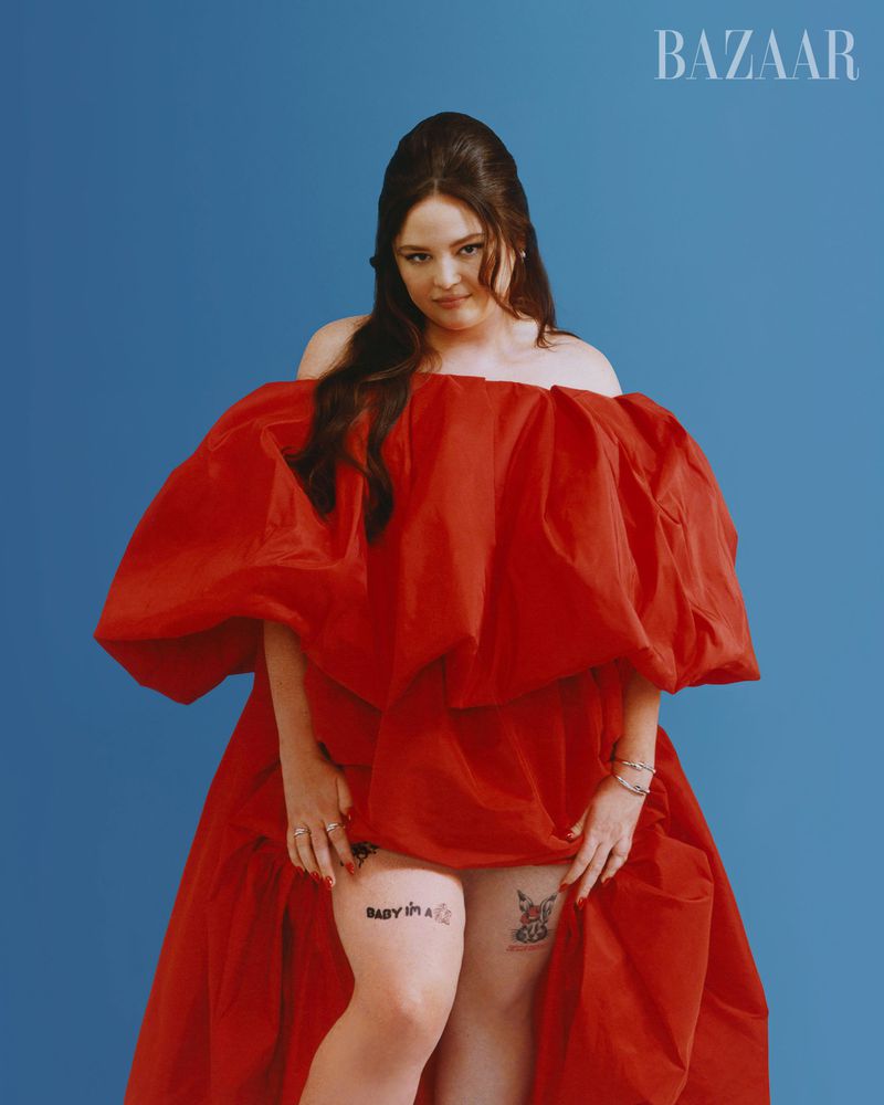 A woman in a red dress poses against a blue background, with 'BAZAAR' visible in the upper right, fashion editorial, studio shot.