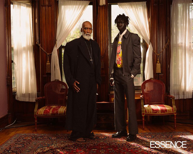 Two Black men in a room with dark wood panelling and large windows, one in a long black coat and one in a grey suit and colorful tie.