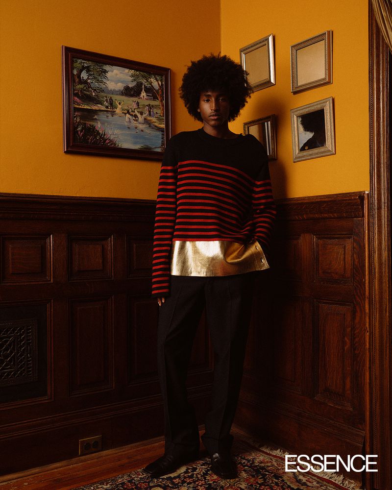 A person with an afro stands indoors with a hand in their pocket, wearing a horizontally striped shirt with a gold panel.