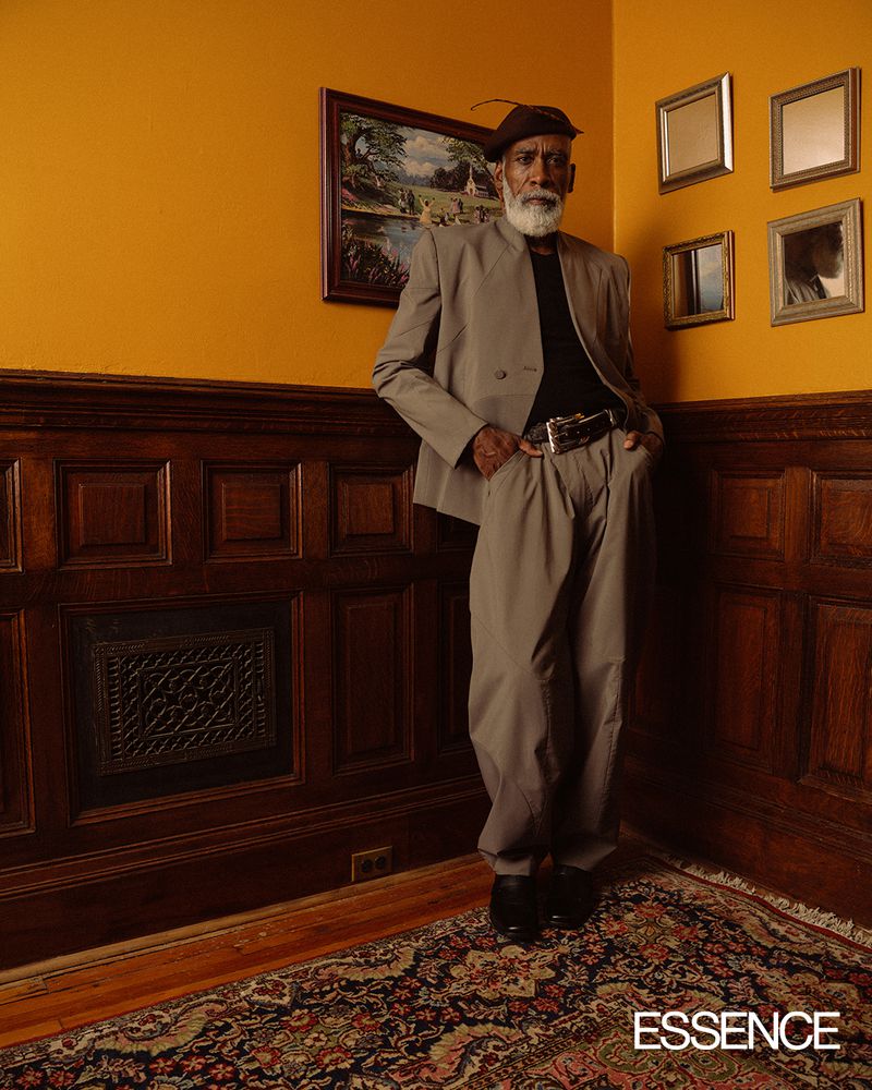 Elderly Black man in tan suit posing indoors near paneled wall and ornate rug with portrait and mirror collection on the wall.