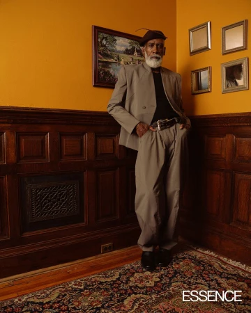 Elderly Black man in tan suit posing indoors near paneled wall and ornate rug with portrait and mirror collection on the wall.