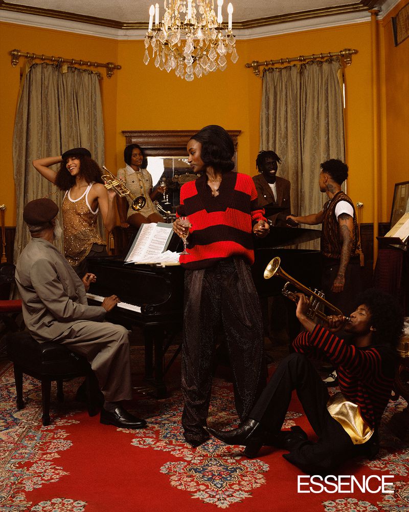 A diverse group of seven people, including musicians playing piano and trumpets, and standing figures, gather in a room with warm lighting.