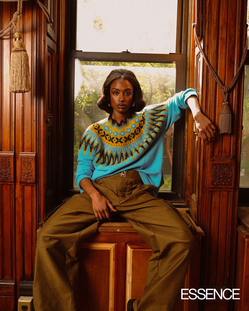 Adult woman poses indoors wearing a patterned, long-sleeved blue sweater and olive-green, high-waisted pants with decorative wood background.
