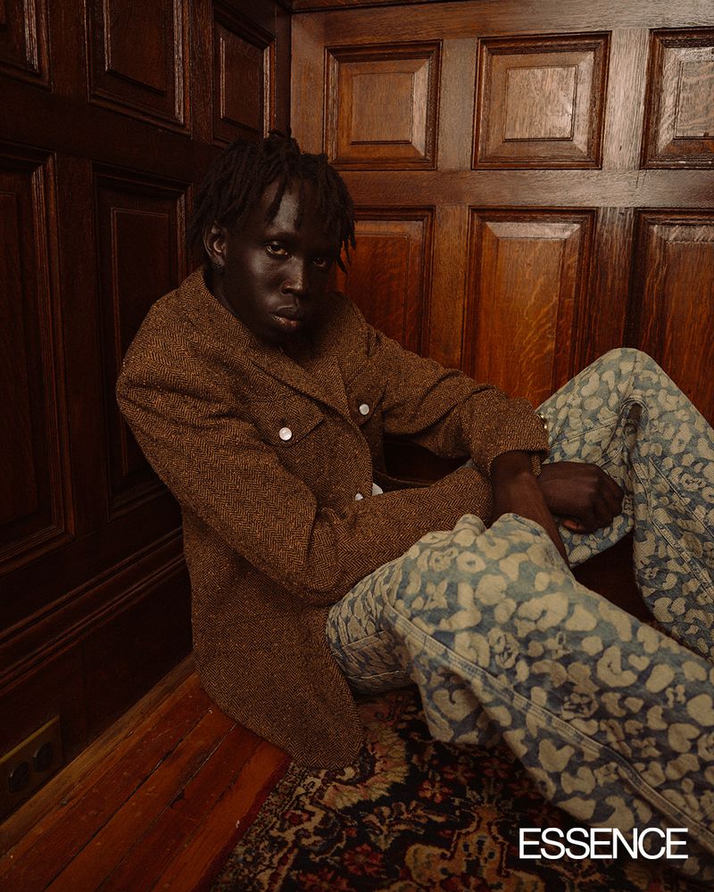 Portrait of an African American man in patterned jeans and brown jacket sitting in a corner near a rug and wood paneling.