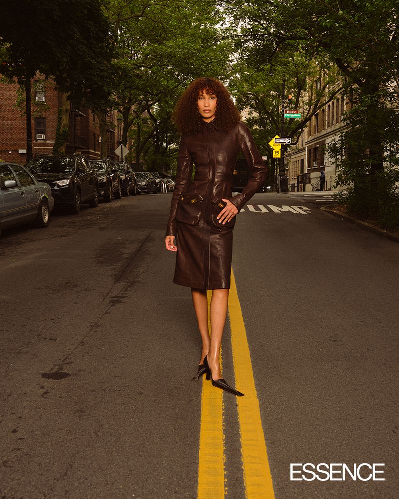 Fashionable woman in leather suit poses on city street with parked cars and trees under overcast sky; Essence logo in corner.