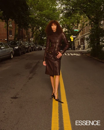 Fashionable woman in leather suit poses on city street with parked cars and trees under overcast sky; Essence logo in corner.