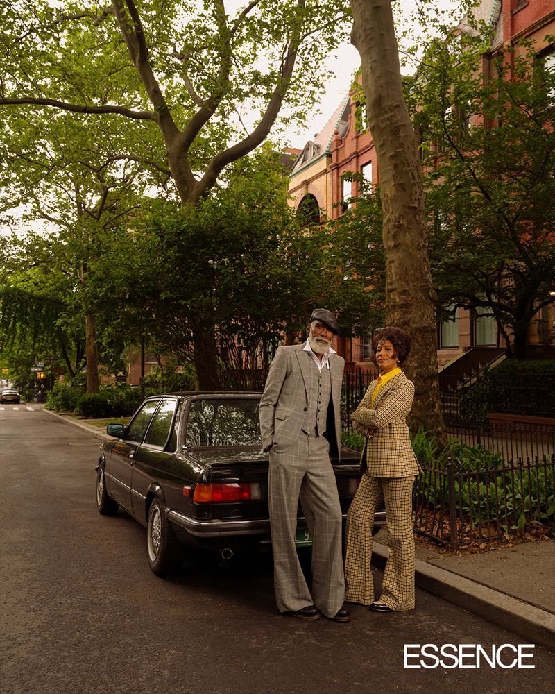 Mature couple poses next to a classic black car on a tree-lined street in front of brick buildings, fashion shoot for Essence.